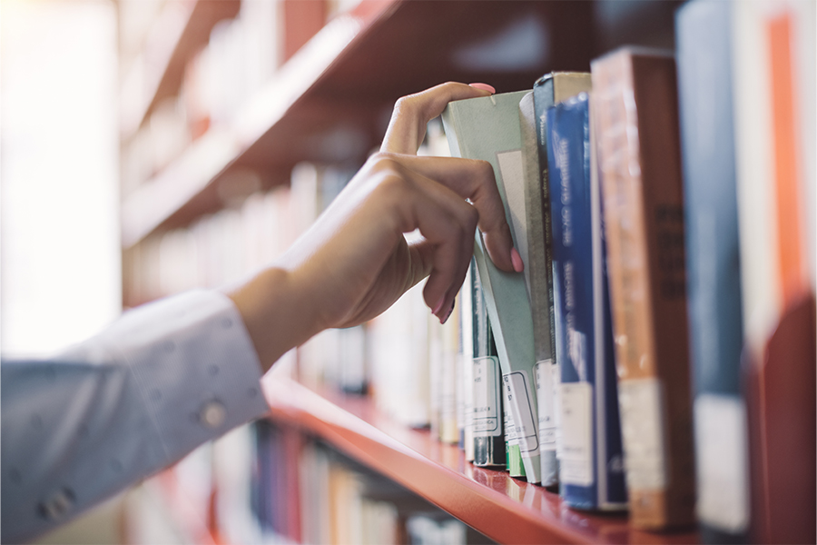 A close-up, shallow-focus shot of a person’s hand with pink manicured nails reaching for a light green book on a library shelf. The background shows blurred rows of books on wooden shelving.