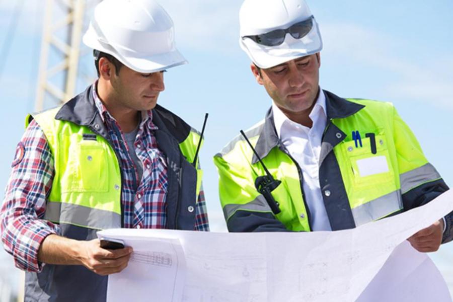 Two civil engineers in hard hats and safety vests reviewing blueprints at a construction site.