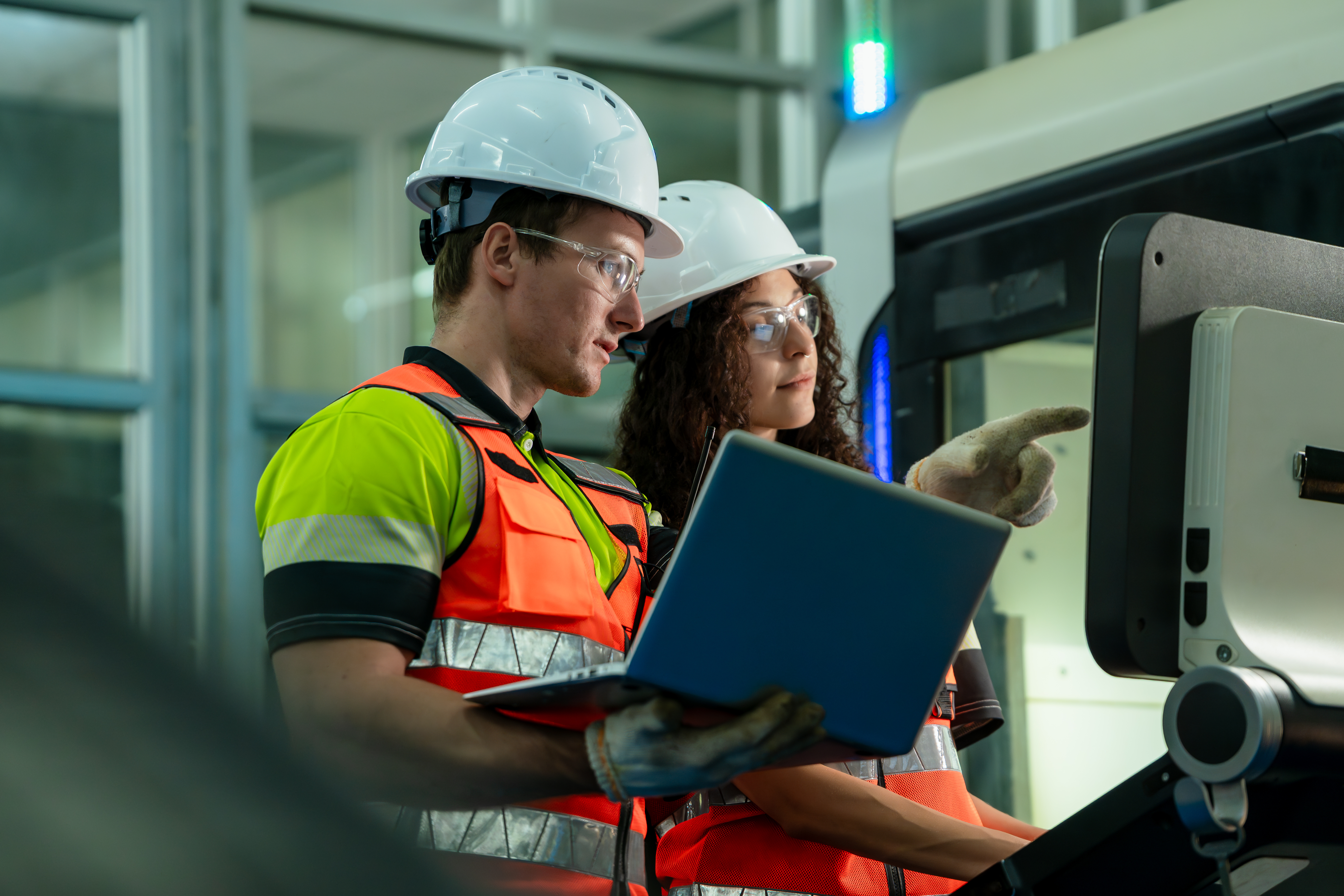 A man and woman with hard hats and safety vests stand looking at a computer screen together. The man points at the computer screen.